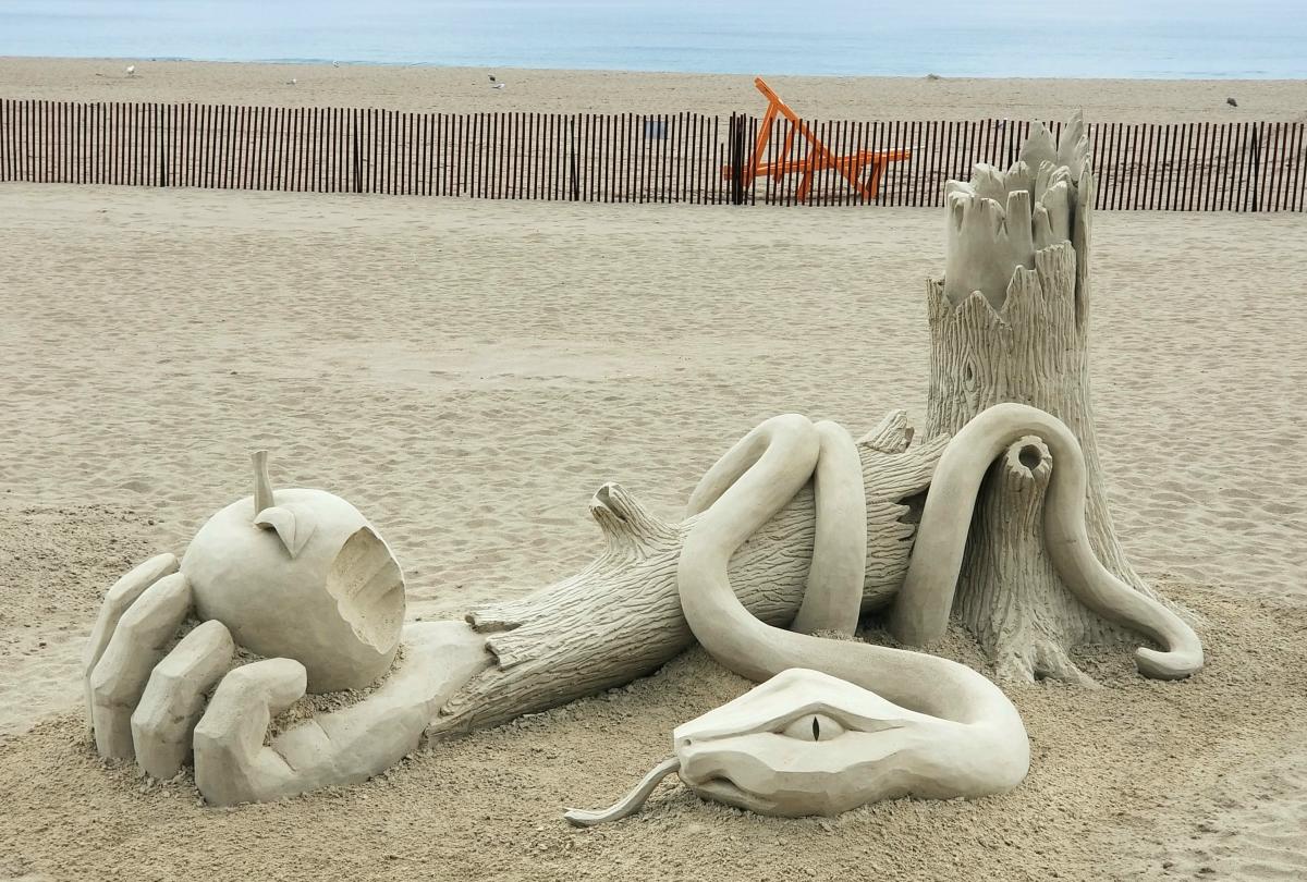 Sand sculpture of a snake slithering over a broken tree trunk that morphs into an arm holding a bitten apple