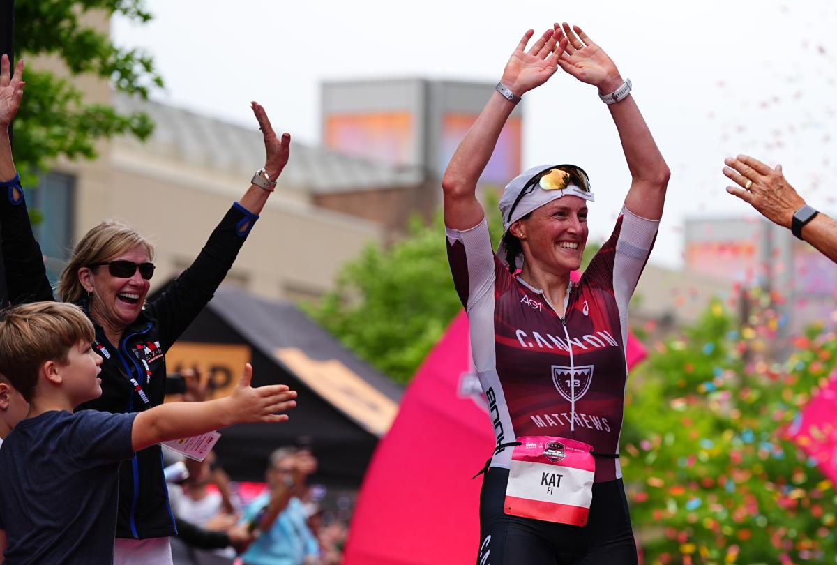 A young white woman in a red and black athletic suit (Kat Matthews) raises her arms in celebration as she crosses the finish line of the Memorial Hermann IRONMAN Texas. Linda Nelson and other crowd members cheer for her as confetti rains down behind them.