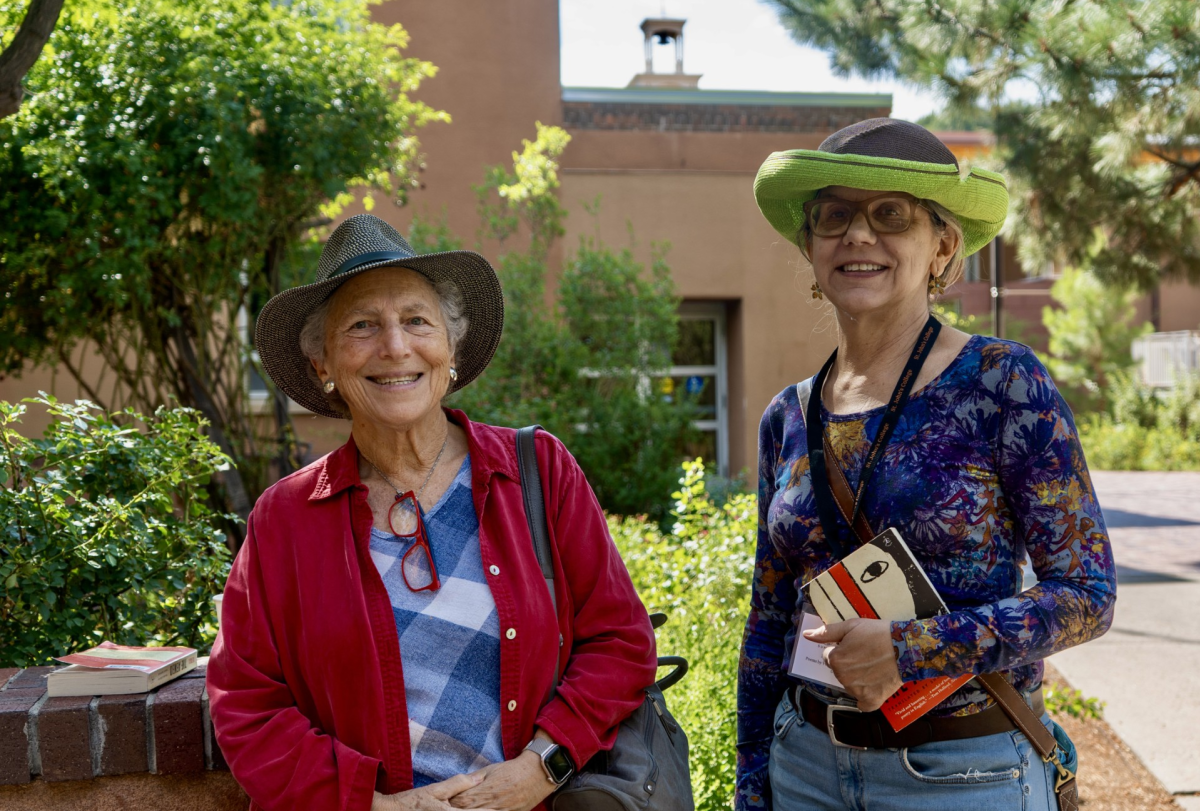 Two smiling women in colorful hats and vibrant clothing stand outdoors, holding books.