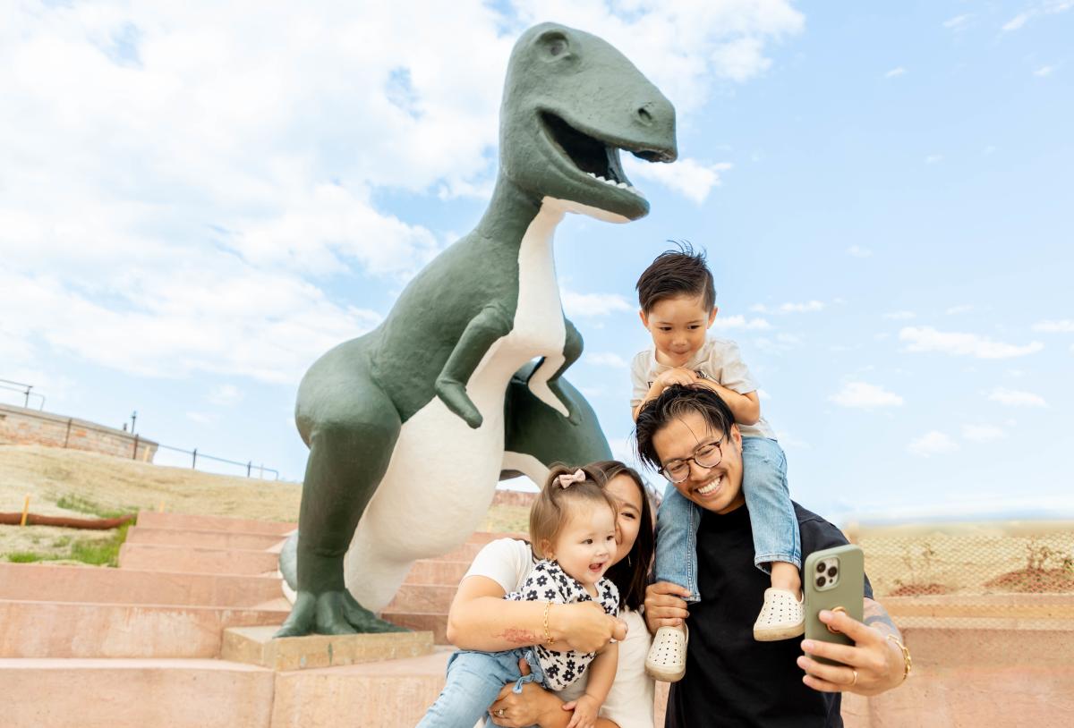A happy family takes a selfie in front of a large green and white dinosaur statue on a sunny day, with smiles and playful expressions.