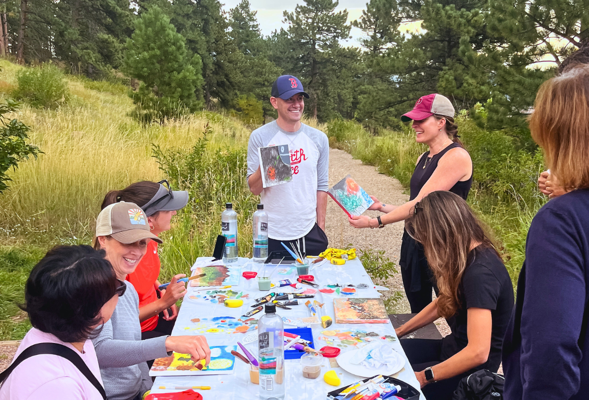 A group sits at an outdoor table off of a trail on open space and shares their painted canvases with one another