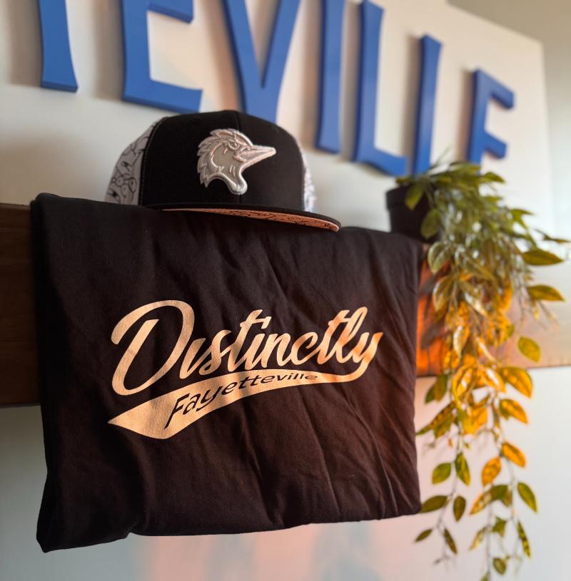 Black “Distinctly Fayetteville” t-shirt with white script lettering and a black-and-white Fayetteville Woodpecker's collectible baseball cap displayed on a shelf, with a green plant beside it. This swag prize is available to win when downloading the free Destination Guide Sept. 10–22.