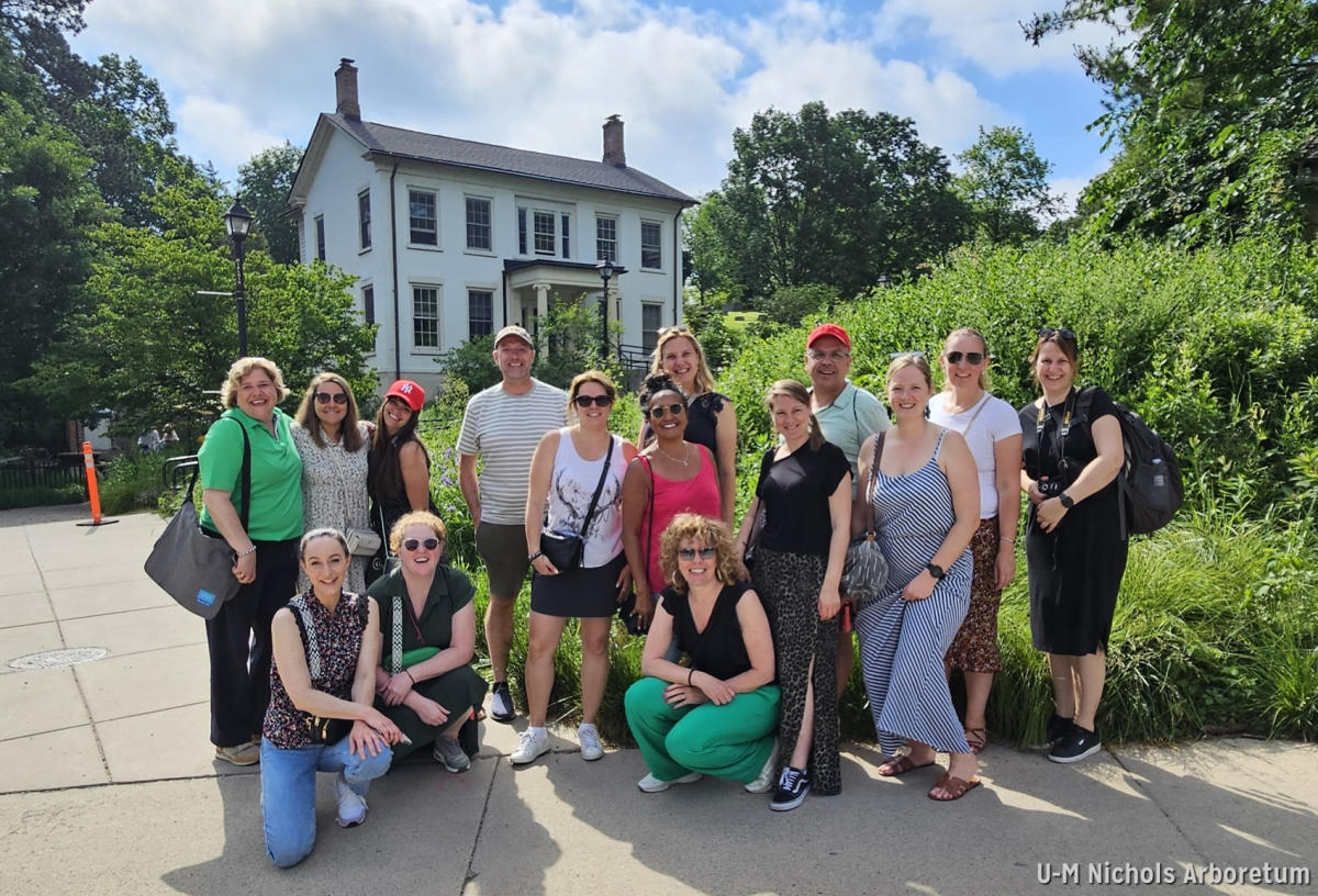 Group poses outdoors in front of historic home surrounded by greenery.