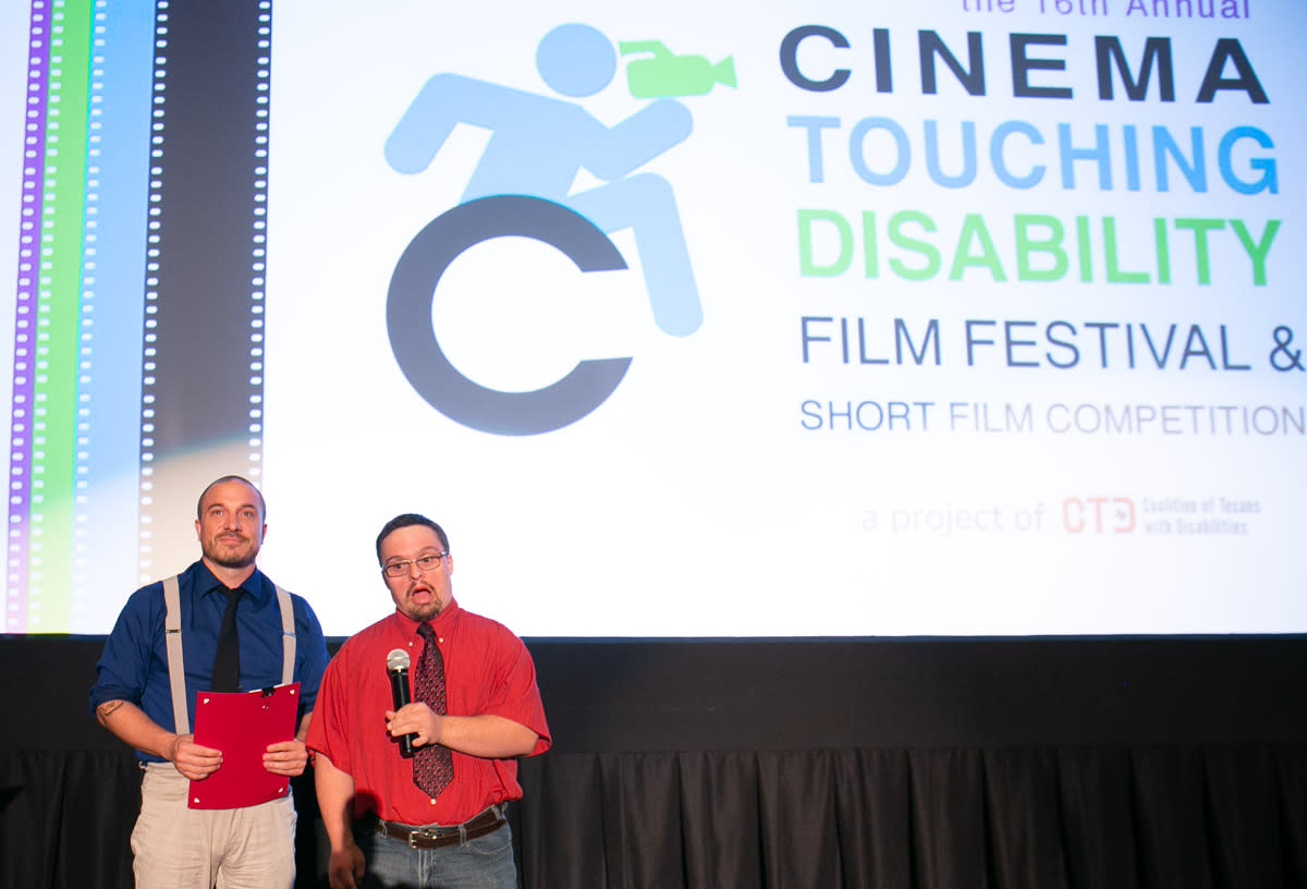 Image of two men standing on a stage speaking with the Cinema Touching Disability logo on a screen behind them.