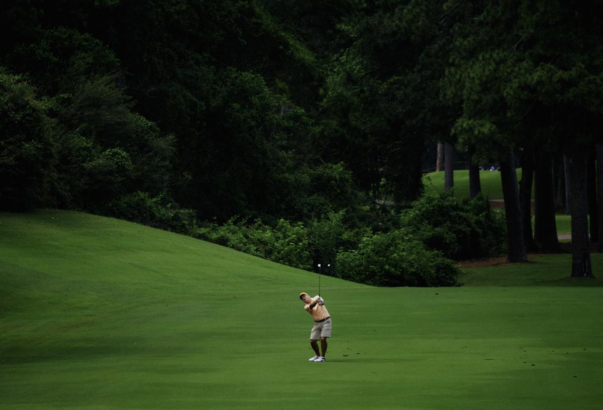 A white man, 50s-60s, swings a golf club to send a golf ball far down the course (behind the camera). He's wearing a peach golf polo and khaki pants, and he's standing at the base of a green hill, behind which is a cluster of trees.