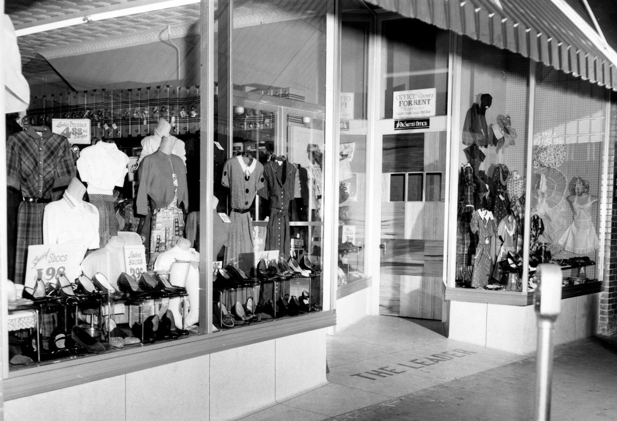 Display windows of The Leader clothing store in Sylva, NC, in the late 1950s, filled with dresses and shoes, with the store name set in the entry floor