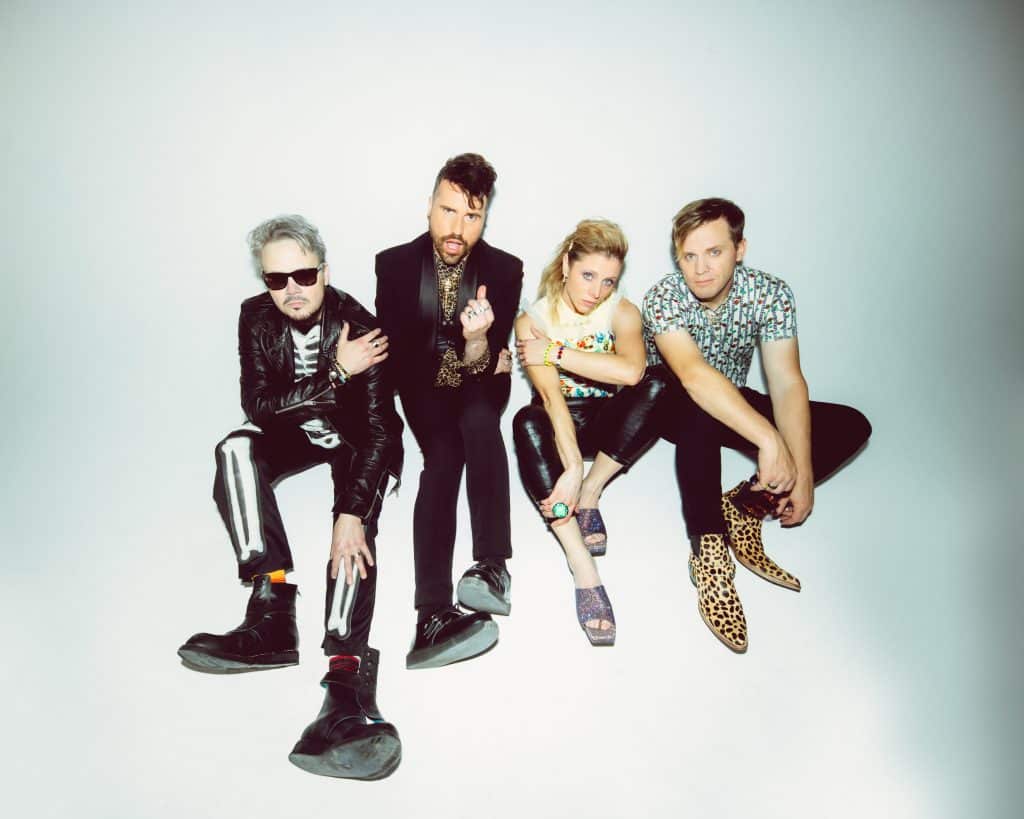 Four members of a music band dressed in stylish and unique outfits posing with playful attitudes against a light background.