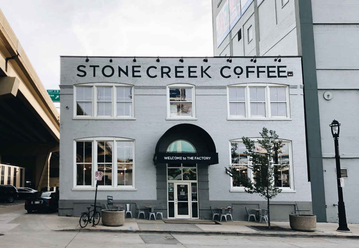 Exterior of Stone Creek Coffee’s white brick building with large front windows, black awning, and outdoor seating along the sidewalk.