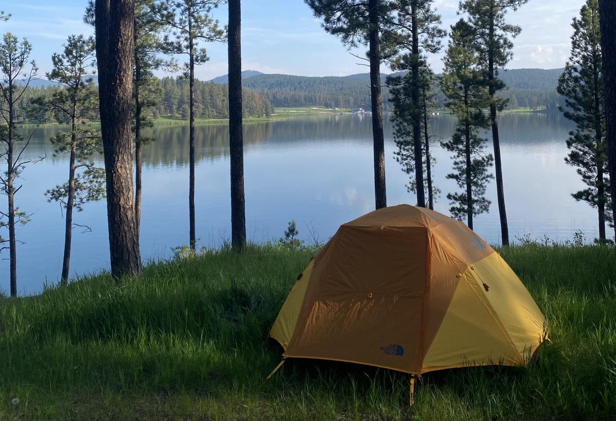 A yellow tent is set in the grass in the shadow of tall ponderosa pine trees under a blue sky overlooking a lake in the Black Hills.