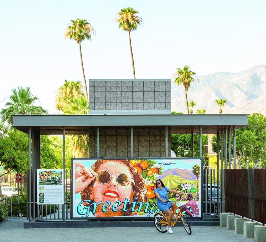  Woman riding a yellow bike in front of a colorful Palm Springs mural, promoting local bike tours amid palm trees and mountain views.