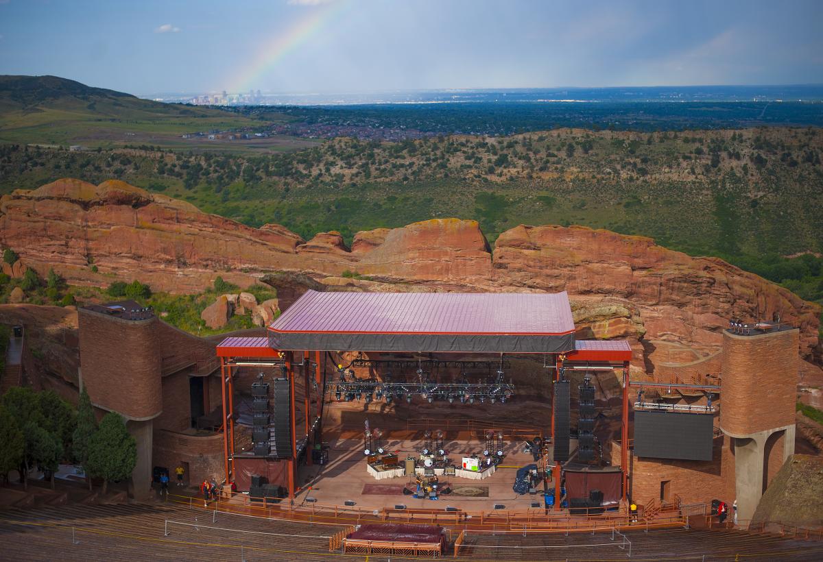 Red Rocks Amphitheatre stage from the open air seats during the daytime with a rainbow and metro Denver in the far distance.