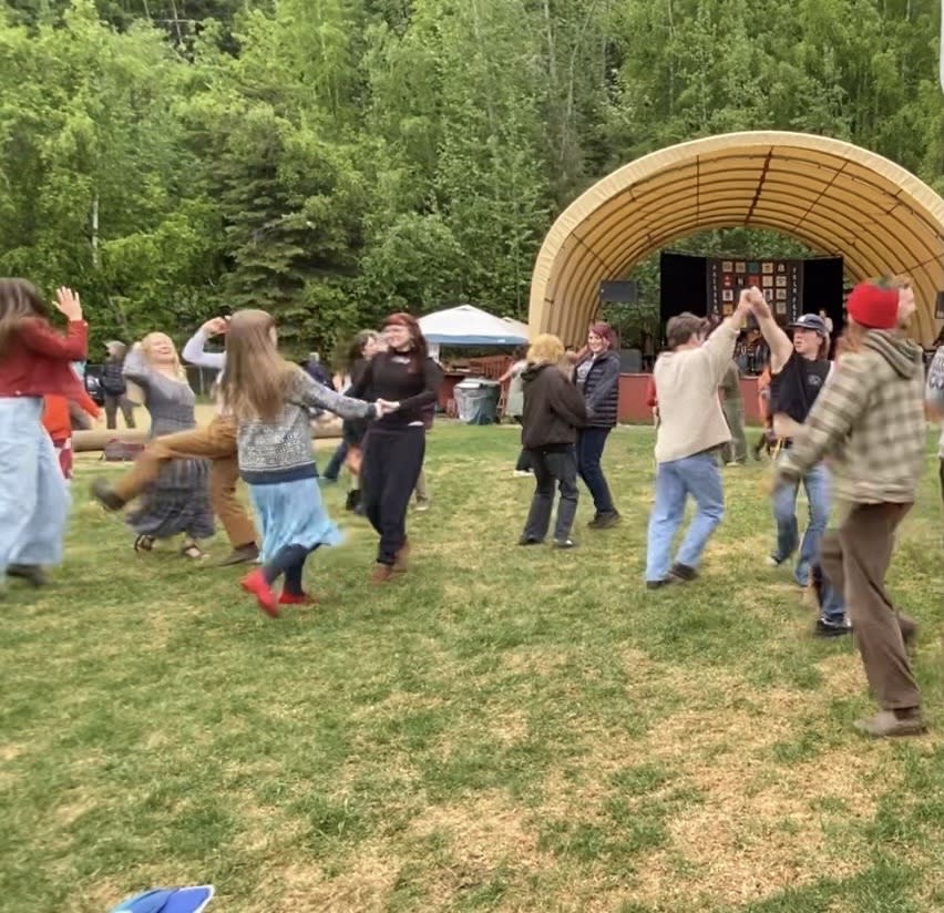 People dancing and celebrating at the Fairbanks Summer Folk Festival on a grassy lawn in front of an outdoor stage surrounded by green trees.