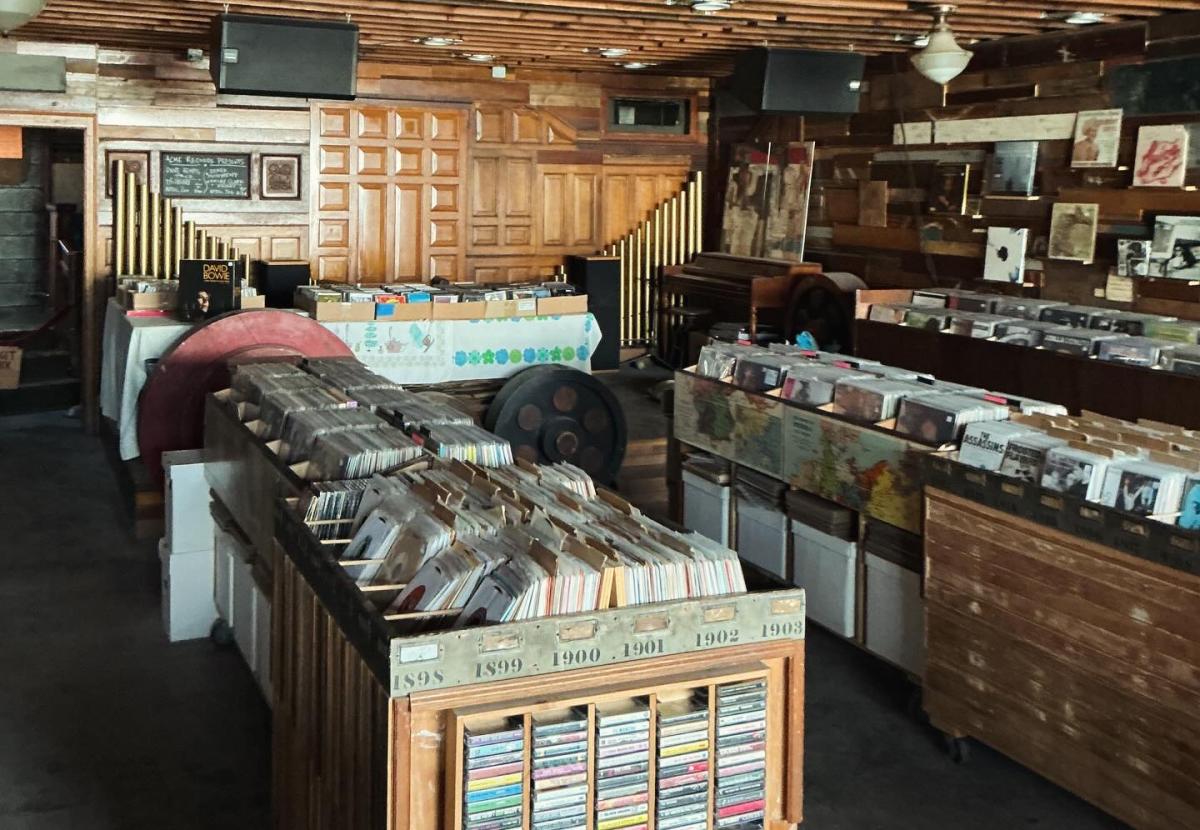 A record shop with a vintage feel features wooden walls, antique fixtures, and rows of vinyl bins filled with albums. Records are organized in large crates and tables throughout the space, with framed artwork and music memorabilia displayed along the walls.