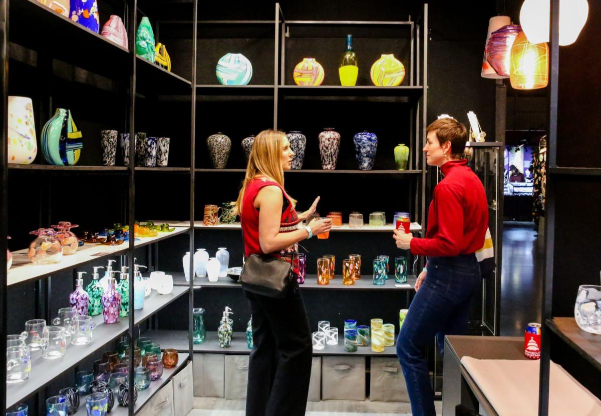 Two women in their early 30s talking in a pop-up shopping display of blown glass works.