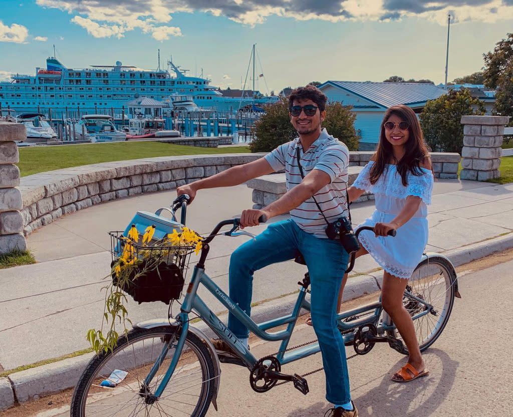 A young couple rides a tandem bicycle past the Mackinac Island harbor with a cruise ship in the background