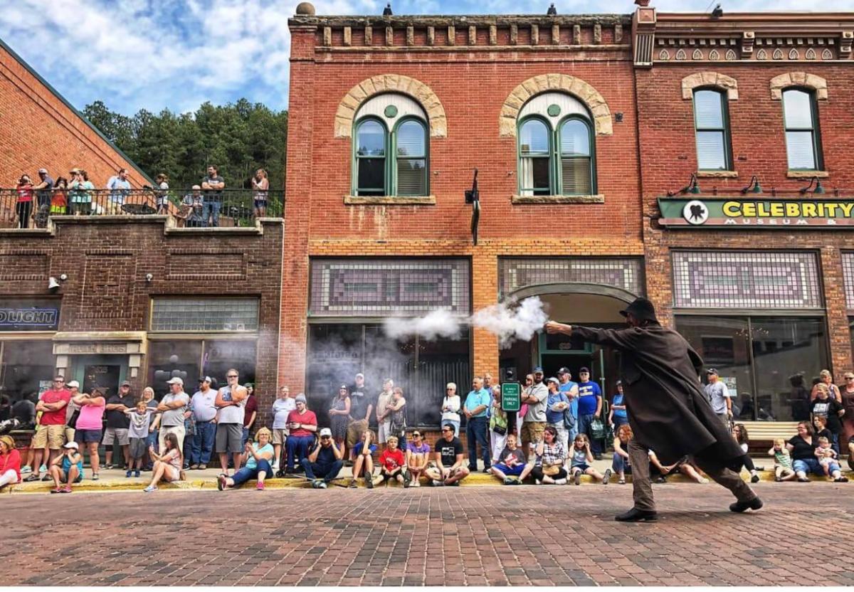A reenactor dressed in replica late-1880s apparel fires his gun on a cobblestone street as visitors, some with their phones out for pictures, look on from the curb.