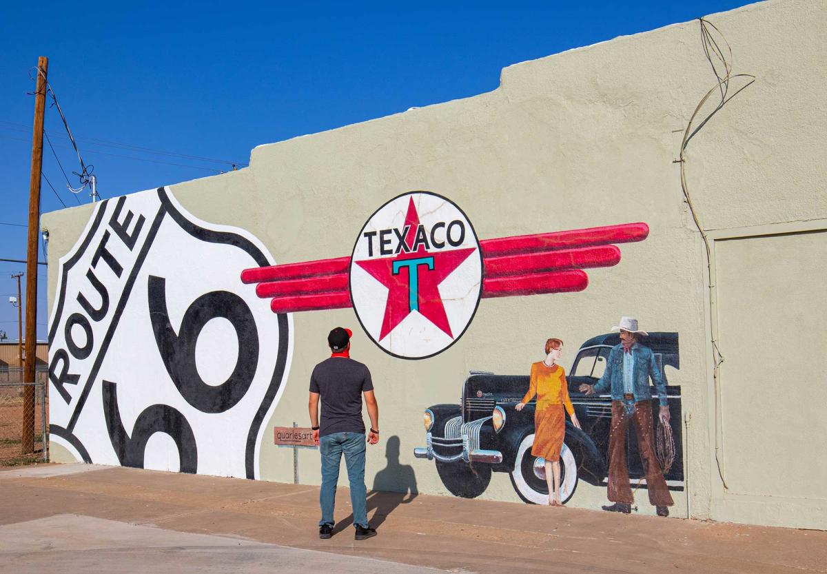 A man stands in front of a mural on a wall depicting two people standing in front of a classic American car with a red and white Texaco logo and a Route 66 sign.