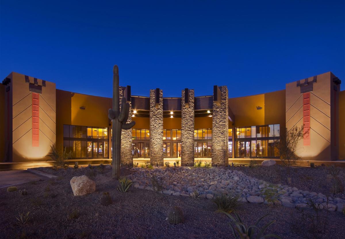 Exterior of Tucson's Desert Diamond Casino. A long, curved building with desert landscaping lit with golden glowing lights against dark night sky
