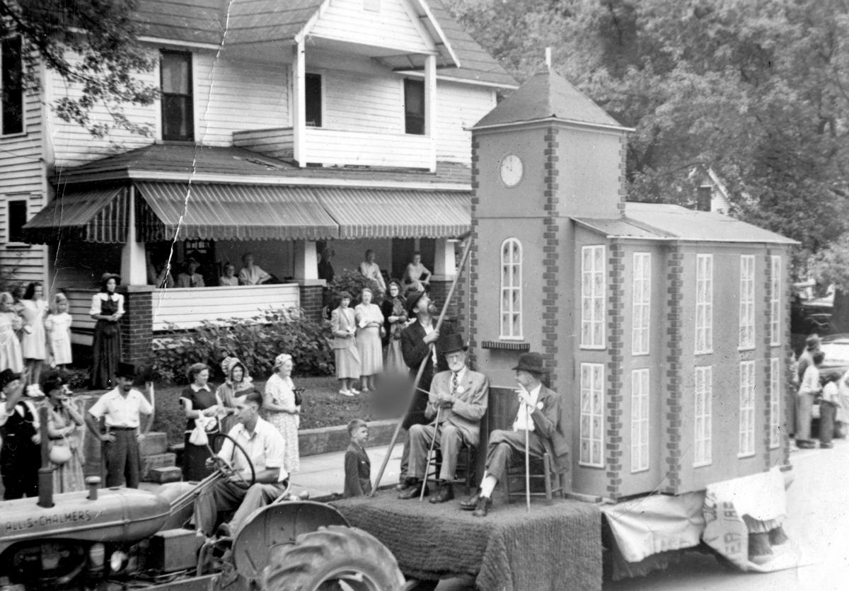 Tractor pulling a float with a replica of the old Webster courthouse and men in top hats past onlookers on Main Street in Sylva, NC, in 1951
