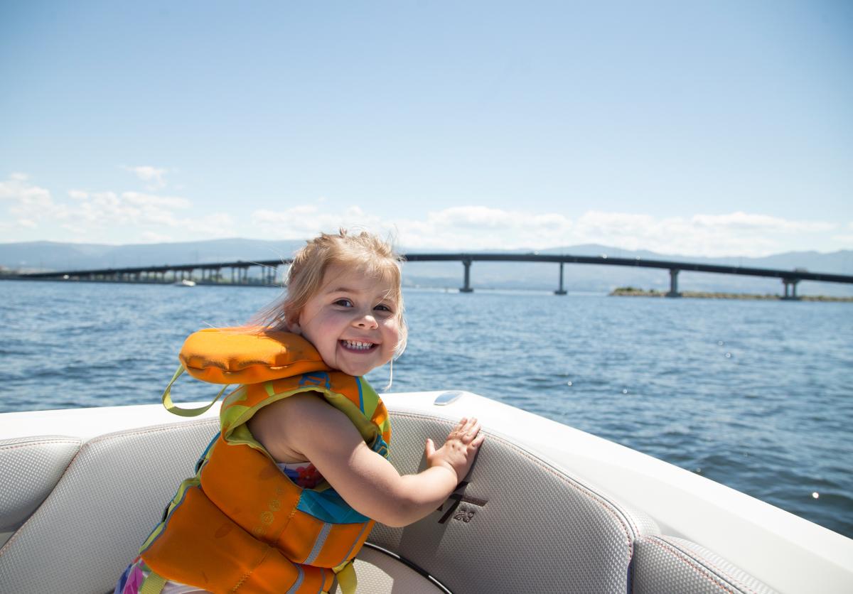 Young girl wearing a life jacket smiling while sitting on a boat, with the William R. Bennett Bridge and Okanagan Lake in the background on a sunny day