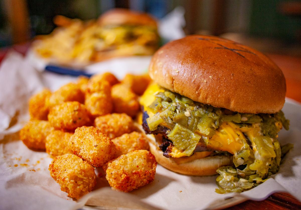 A close-up of a cheeseburger with melted cheese and green chiles, beside a serving of tater tots on a paper-lined tray. Another similar burger is slightly visible in the background.