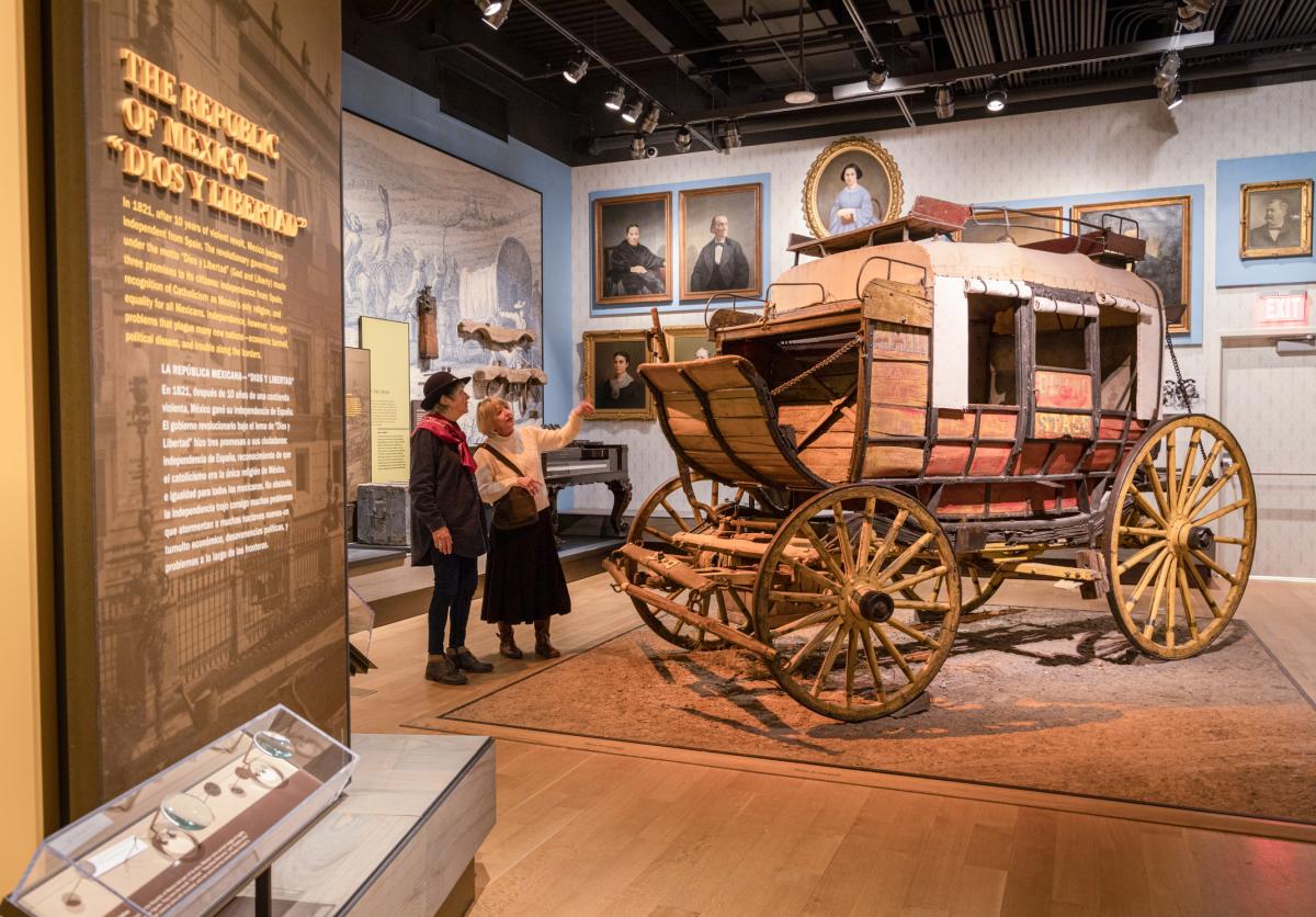 The image shows a museum exhibit featuring a historic stagecoach. Two individuals stand nearby, observing the display. The room is decorated with paintings and photographs.
