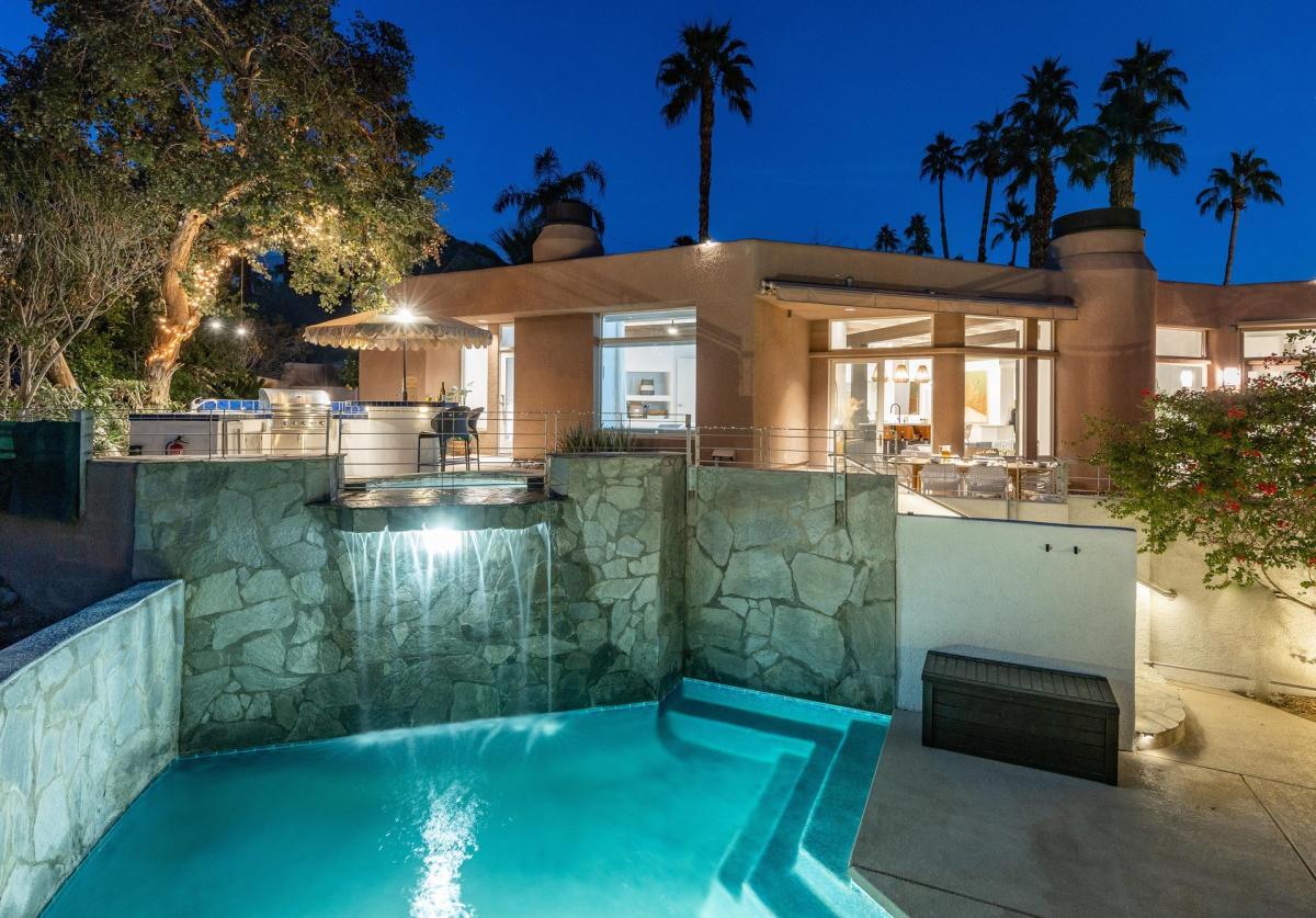 An illuminated outdoor swimming pool at twilight with a waterfall feature, adjacent to a luxurious villa with outdoor lighting and palm trees in the background.