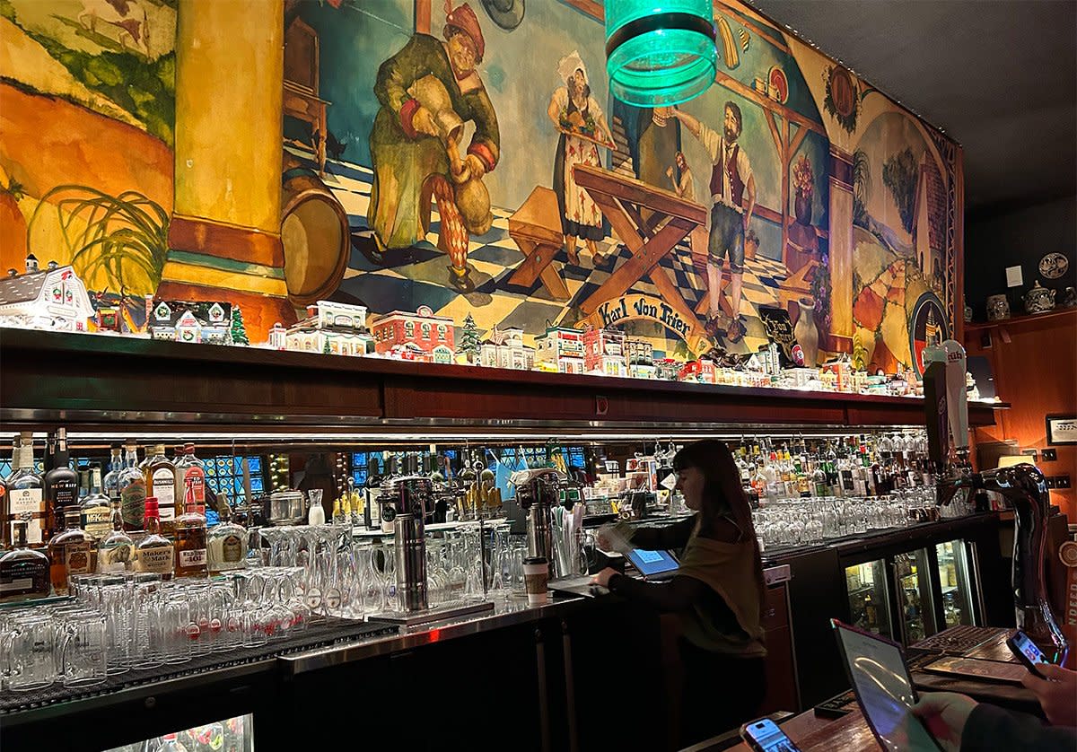 A bartender works behind a bustling bar lined with glassware and taps, set beneath a colorful, German-inspired mural that brings a lively, old-world atmosphere to the space.