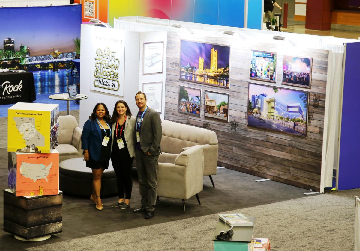 Three smiling team members in a trade show booth surrounded by photos of Sacramento landmarks