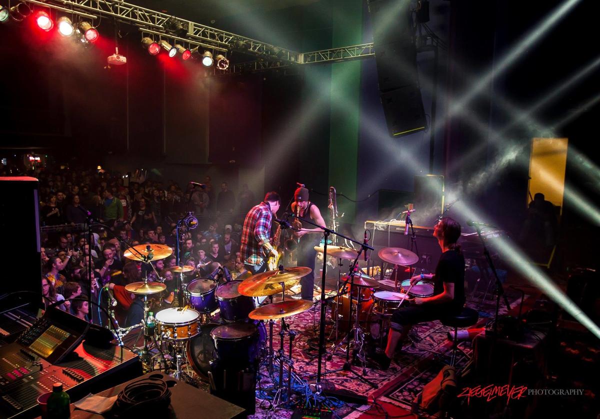 Image is of a backstage view of artist playing their music at the Madison Theater with fans off the stage.