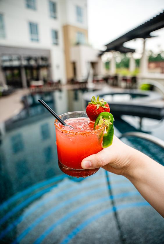 A young woman holds a bright red drink over the bright blue pool at Hyatt Centric. The drink is rimmed with Tajin, a whole strawberry, and a jalapeno slice.