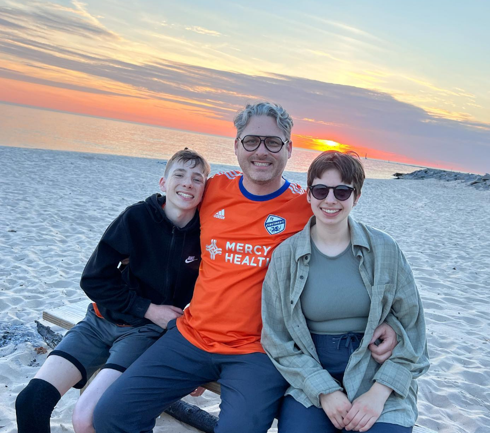 son, father and daughter sit on bench bench. they are on the beach with the sun setting over lake michigan behind them