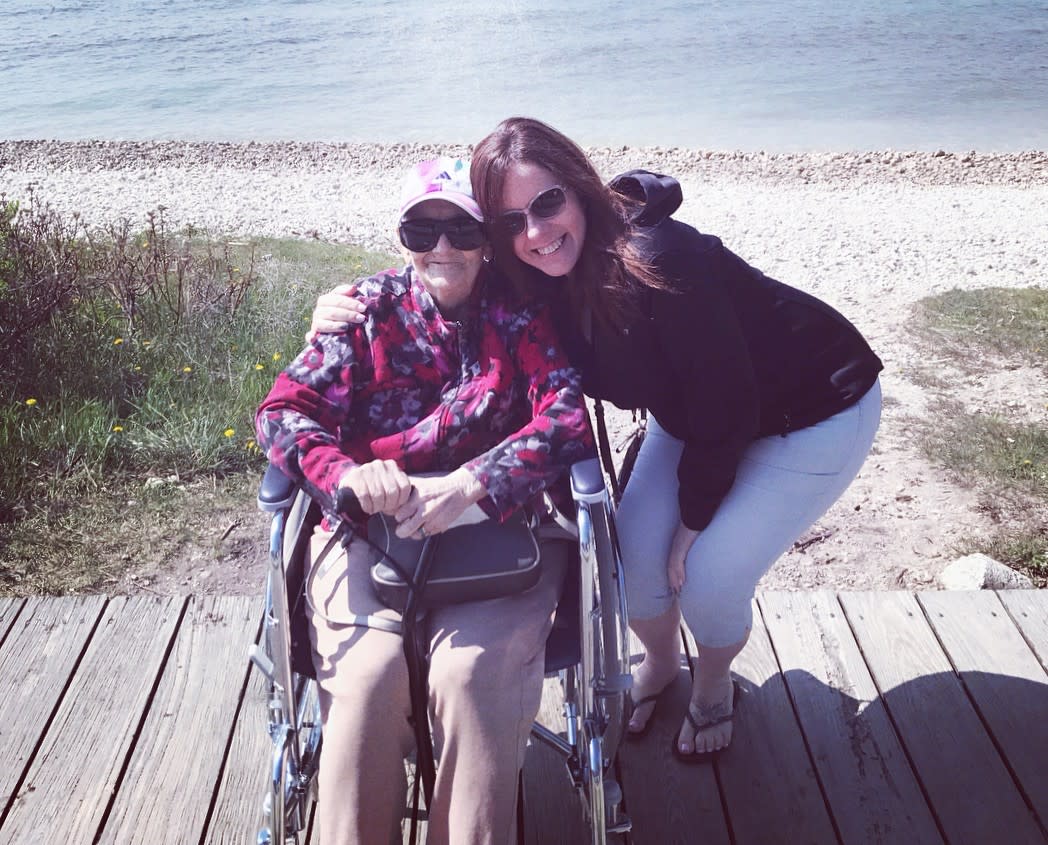 A younger woman hugs an older woman sitting in an electric wheelchair near the water on Mackinac Island