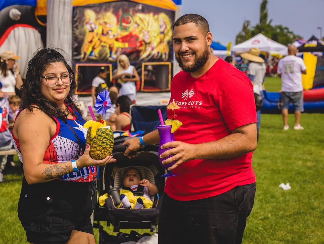 A smiling couple at an outdoor festival holds colorful tropical drinks, one served in a hollowed pineapple and the other in a purple cup with fruit garnishes. Their baby sits in a stroller between them, while other festivalgoers and inflatable attractions are visible in the background.