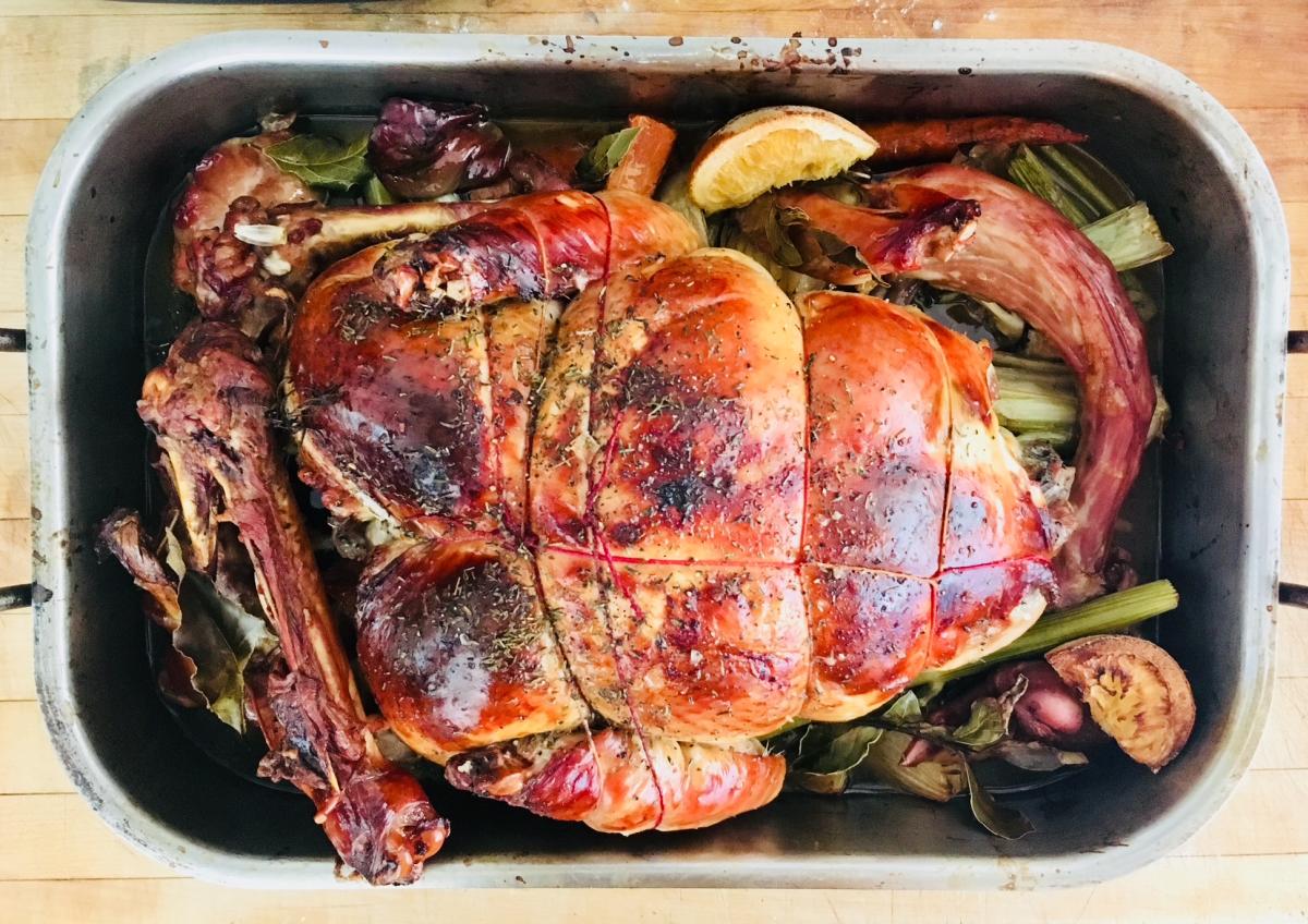 A fully roasted turkey sits in a large metal roasting pan, its skin browned and glossy. The turkey is tied with kitchen twine and surrounded by roasted vegetables, herbs, and citrus halves.