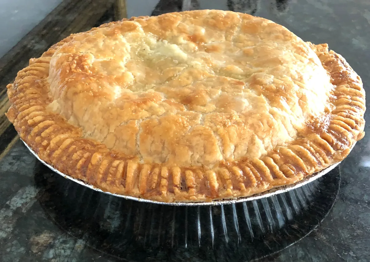 Golden brown pot pie with a flaky, crimped crust baked in a round metal pie tin and resting on a dark countertop.