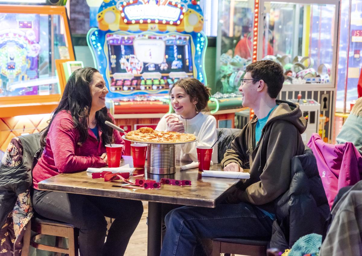 Family of three sitting around a table eating pizza with arcade games in the background at Press Start in Rapid City, SD