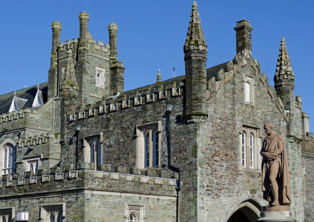 A historic stone castle with crenellated towers against a clear blue sky. A prominent statue of a robed figure stands in the foreground, conveying grandeur and history.