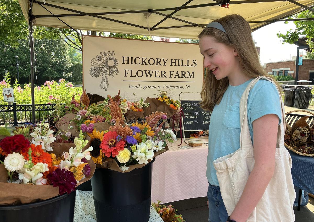 A young woman wearing a light blue shirt is carrying a white shopping bag. She is looking a flowers at a market stall.