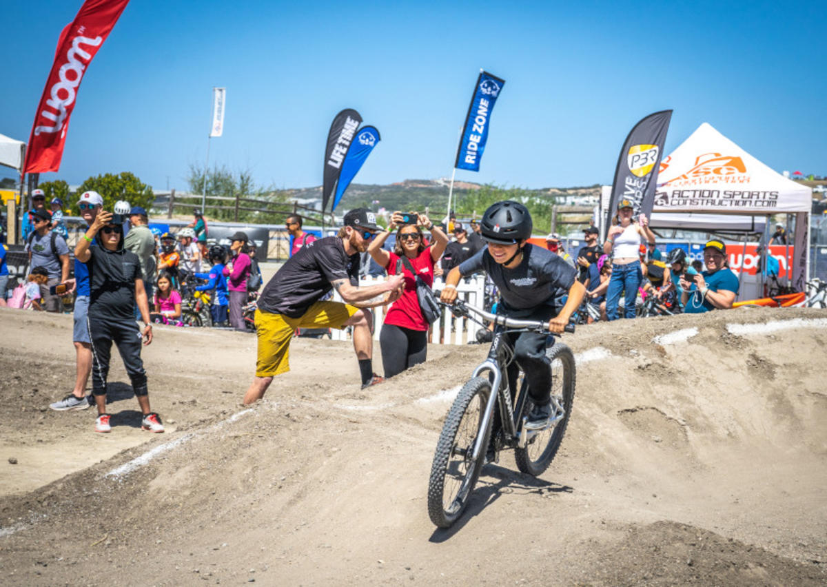 This is an image of a teen boy riding on a bicycle with a helmet on the dirt hills at Sea Otter Classic cycling event in Monterey, California. THere are masses of people gathered in the background at various expo tents