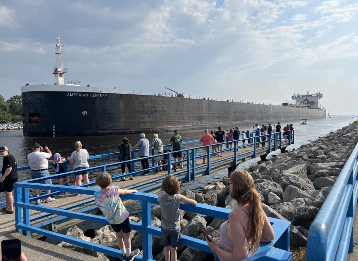 crowd stands along blue railing of muskegon channel watching the freighter american century sail through