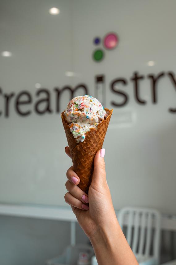 A young woman with light pink nails holds up a waffle cone filled with what appears to be birthday cake flavored ice cream (white ice cream with rainbow sprinkles mixed in). The background (out of focus) features the "Creamistry" name, where there "i" looks like a chemistry test tube with colorful bubbles floating above it.