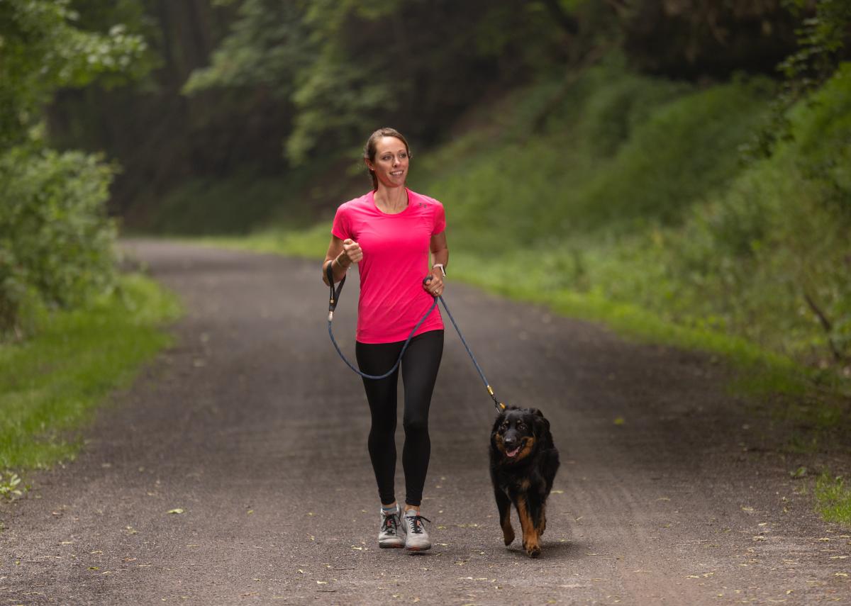 A woman in a bright pink shirt and black pants walks a black and brown, medium-sized dog on a gravel through a wooded area.