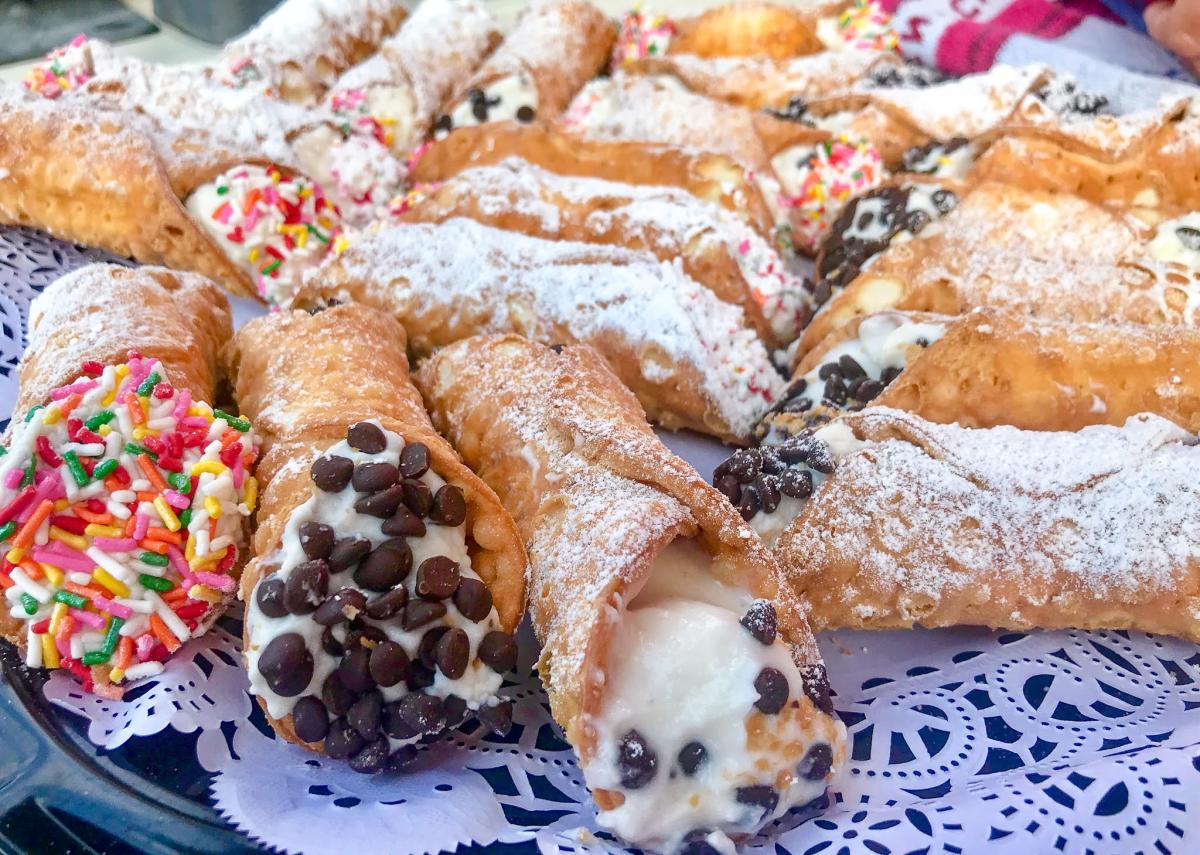 Image is of different cannoli's that are golden brown, dusted in powder sugar, with toppings like chocolate chips and sprinkles.