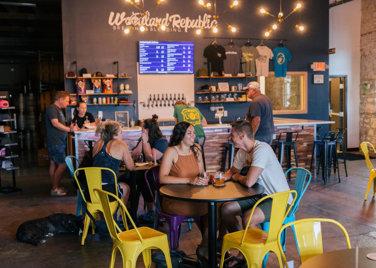 Casual brewery scene with patrons chatting at tables. Warm lighting, colorful chairs, bar backdrop with taps and apparel display, creating a lively, relaxed ambiance.
