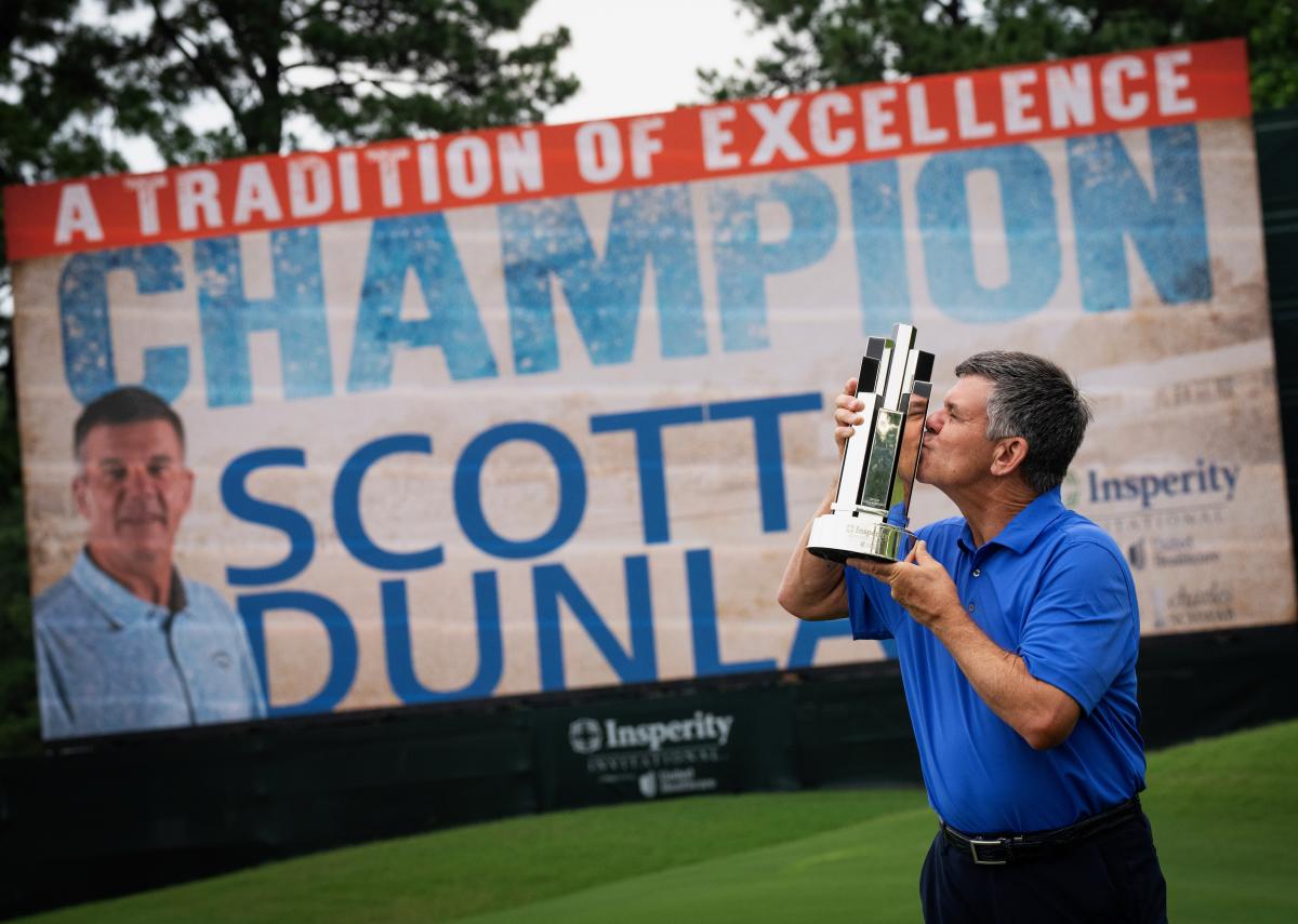 A white man, 50s-60s, holds up and kisses his Insperity championship trophy. He is wearing a blue sports polo and black slacks. The trophy looks like a ring of six mirrored pillars rising from the base. Behind Dunlap and his trophy is a larger-than-life banner featuring his face and the words, "A TRADITION OF EXCELLENCE" (in white text on a red background), "CHAMPION SCOTT DUNLAP" (in blue text on a white background), and the "Insperity Championship" title and logo.