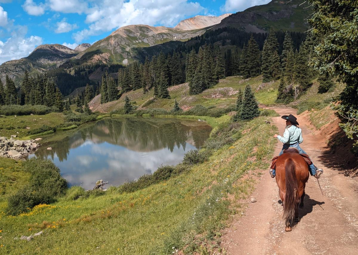 A cowgirl in jeans and a light blue button up on horseback looking at the lake and mountains in the distance while traveling down a dirt road in the San Juan Mountains.
