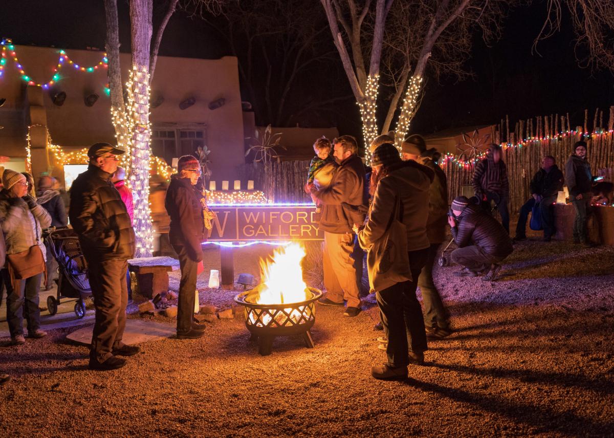A group of people gathered around a fire pit in an outdoor space decorated with colorful string lights. A sign in the background reads "Wilford Gallery.