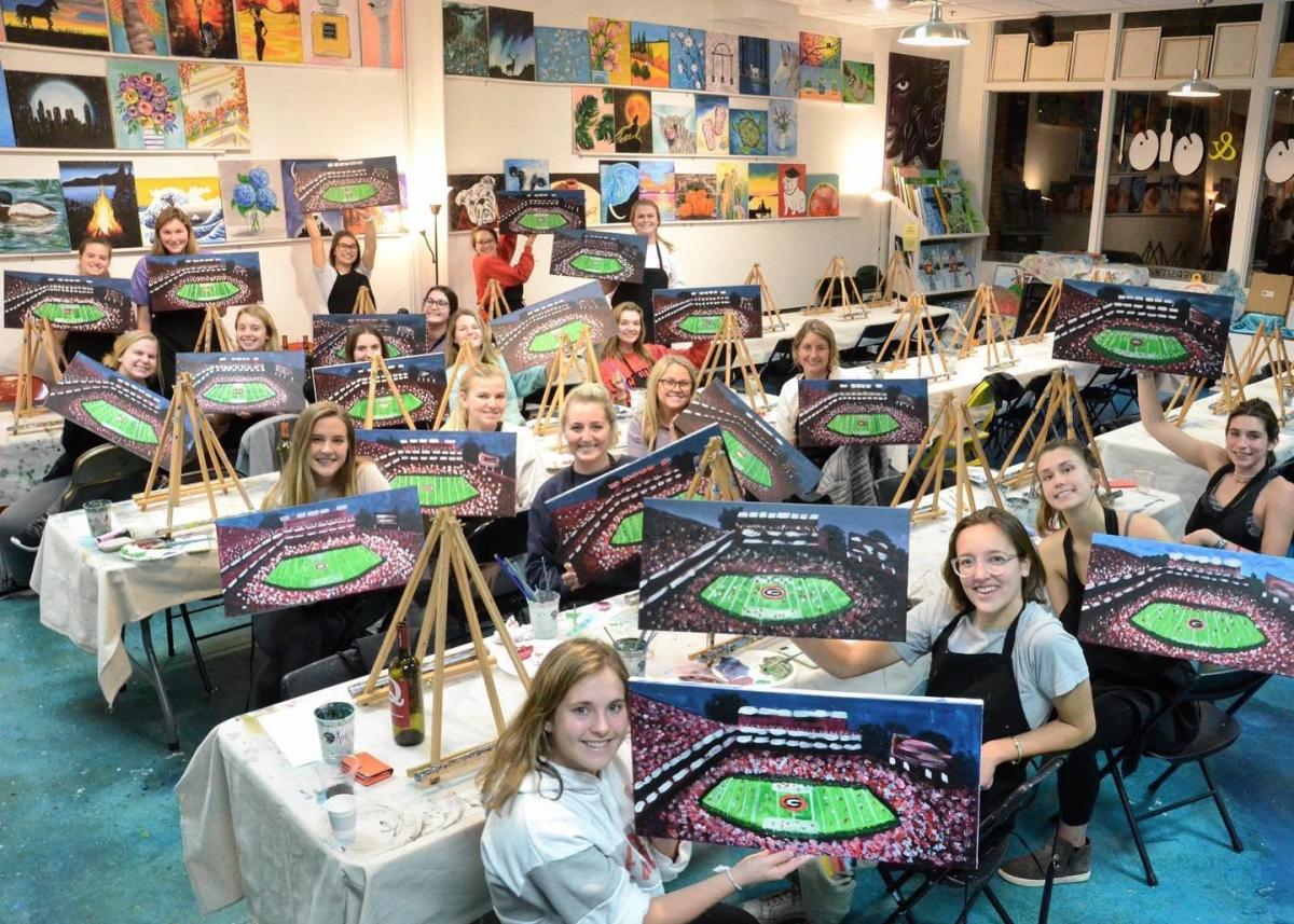 A group of about 20 hold up paintings they did of Sandford Stadium in a class at Artini's