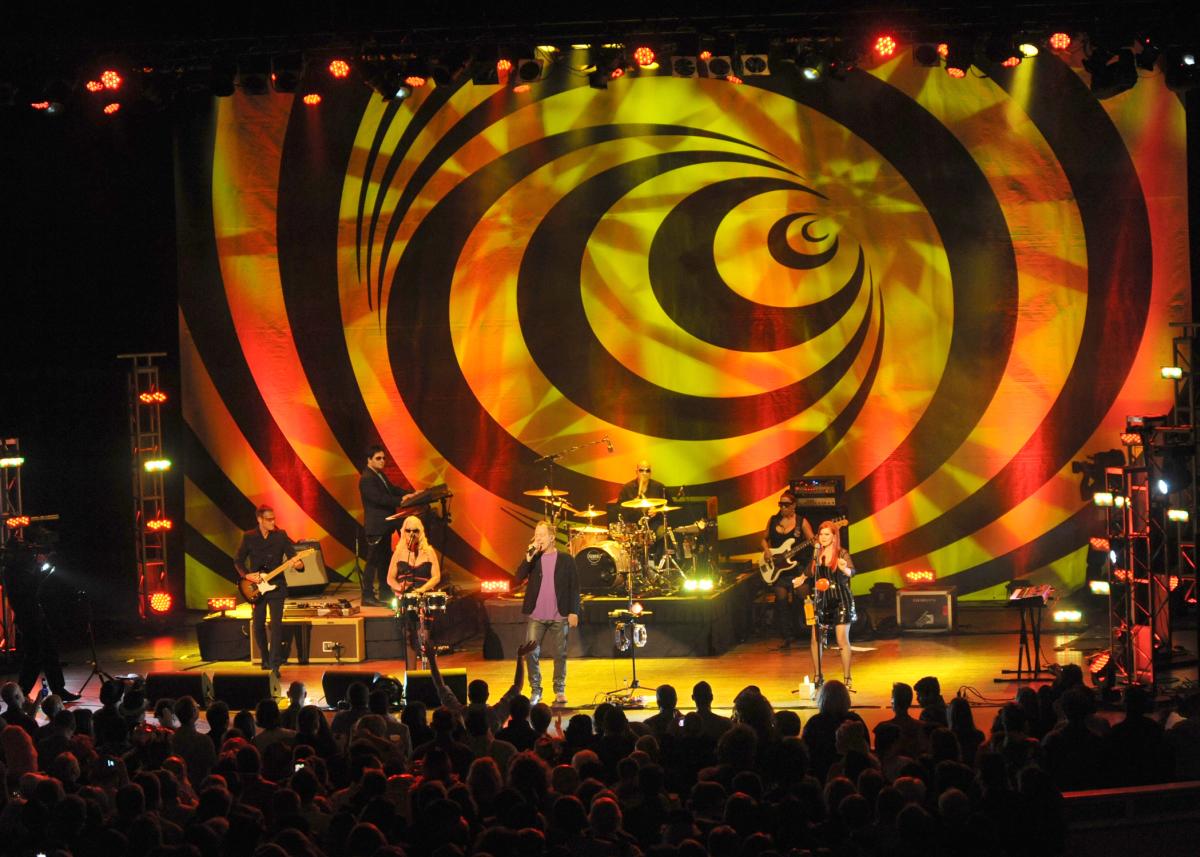 The B-52s perform live at The Classic Center in Athens, with bold yellow-orange striped backdrop seen from the darkened audience below.